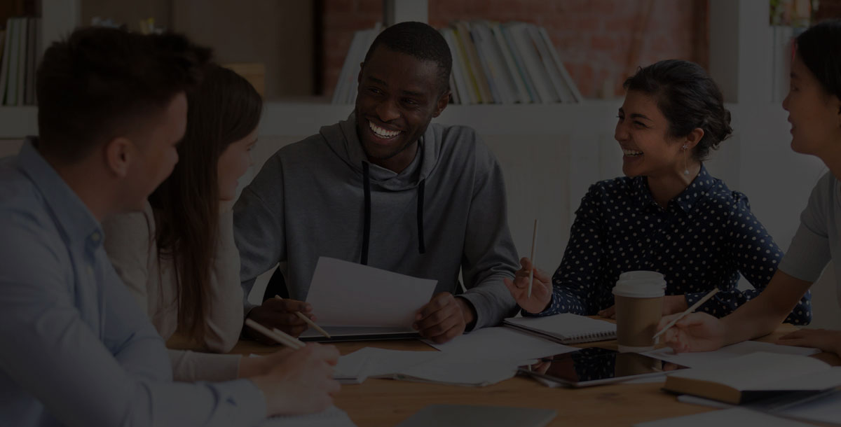 Five people sit at a table, engaged in a friendly discussion, with notebooks and papers in front of them.