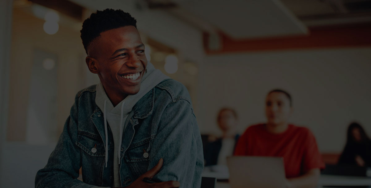 A smiling student in a denim jacket sits in a classroom, with others in the background.