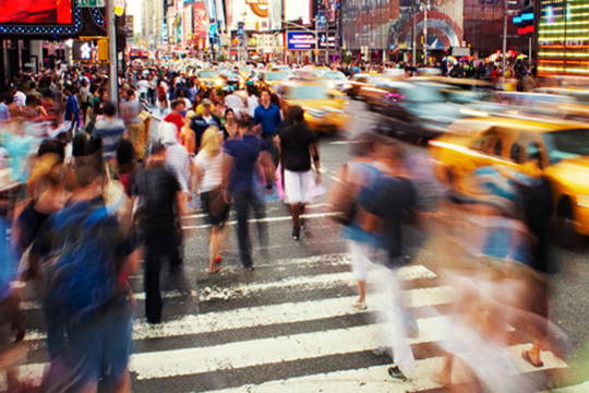 Blurred image of busy city street with people crossing and yellow taxis driving by.